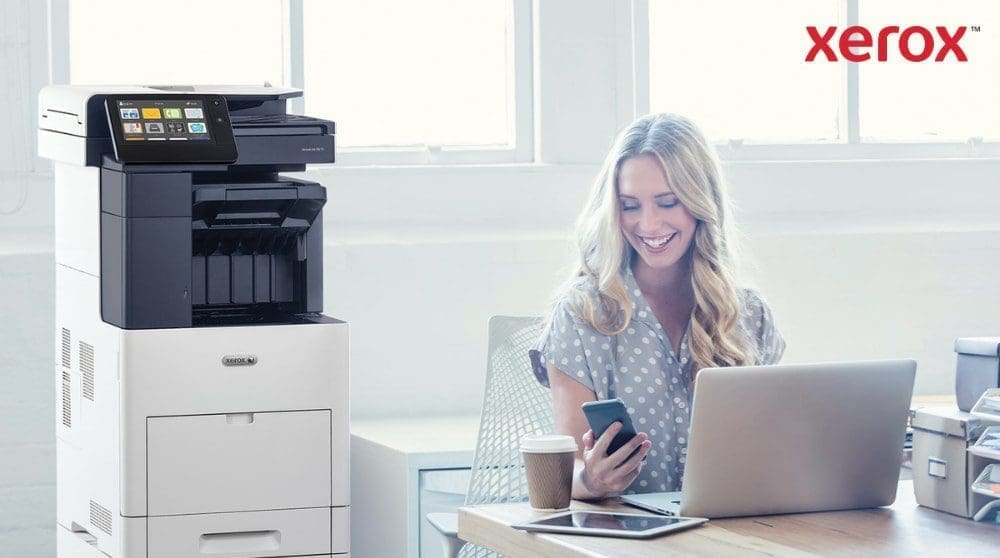 Xerox® ConnectKey® : intégration transparente des flux de travail Une personne regarde joyeusement son smartphone, assise à un bureau avec un ordinateur portable. Une imprimante multifonction Xerox VersaLink B605 est visible à côté du bureau, dans un espace de travail lumineux.